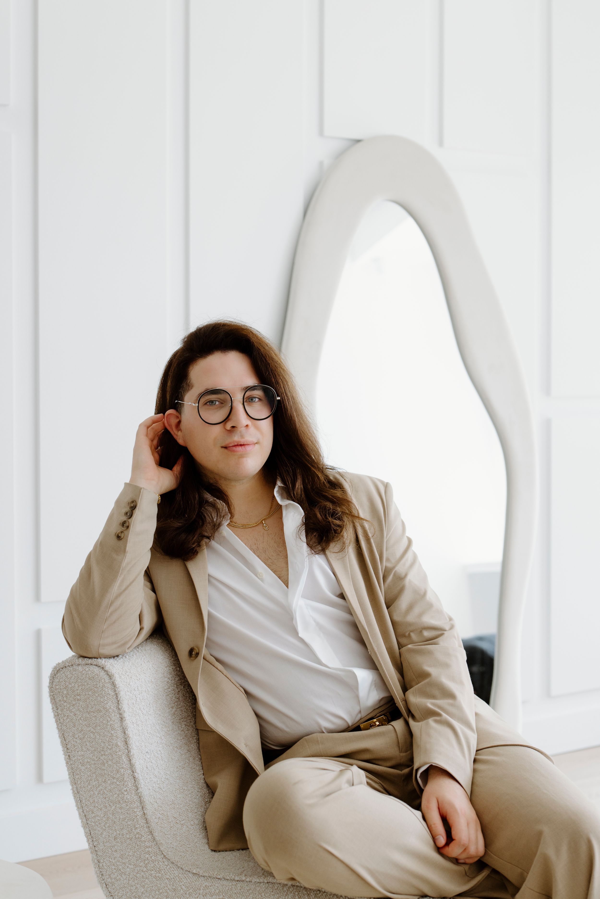Professional wearing a beige suit and glasses sits elegantly in a modern white chair.