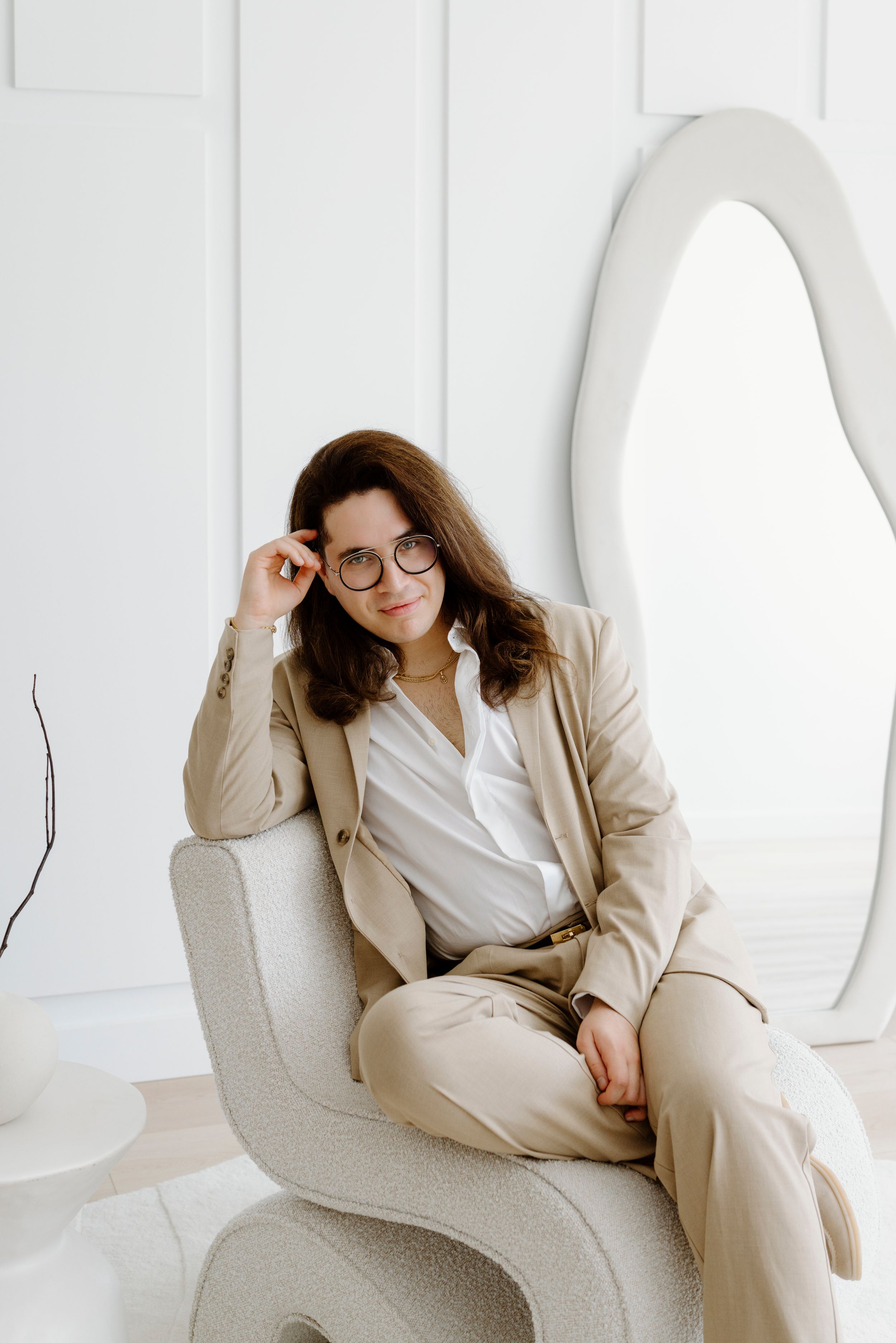 Professional wearing a beige suit and glasses poses elegantly on a white armchair in a minimalist setting.
