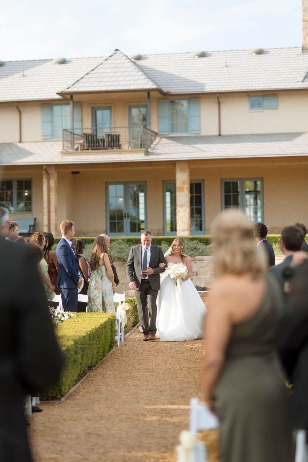Wedding guests gather outside a beige building on a dirt path during an outdoor ceremony.