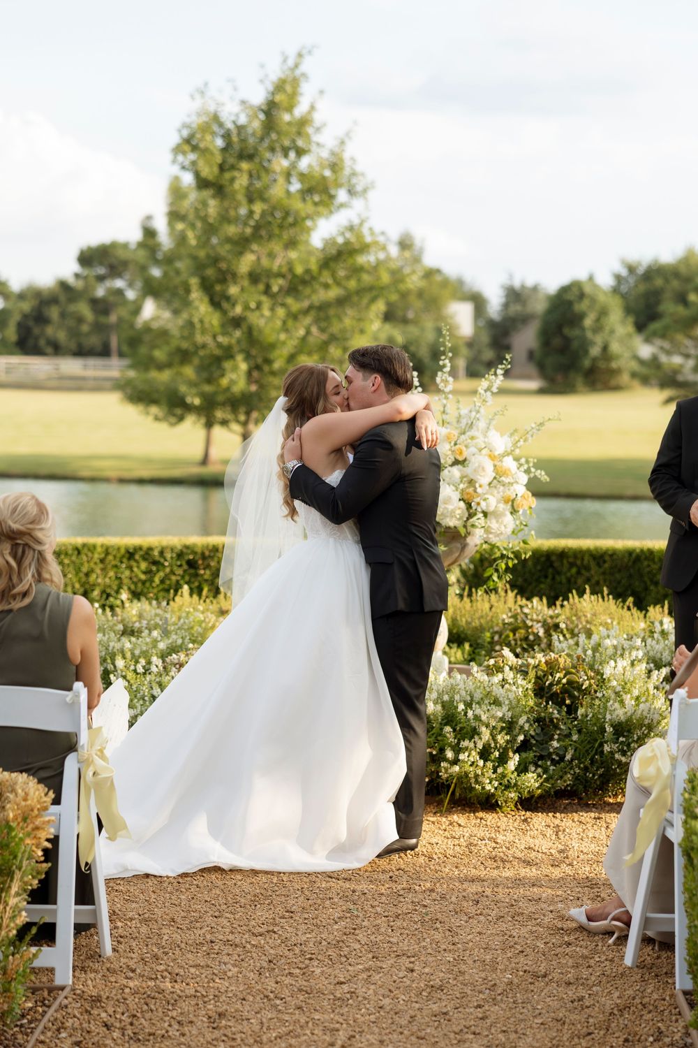 A couple shares a romantic dip kiss during their outdoor wedding ceremony with white chairs and greenery in the background.