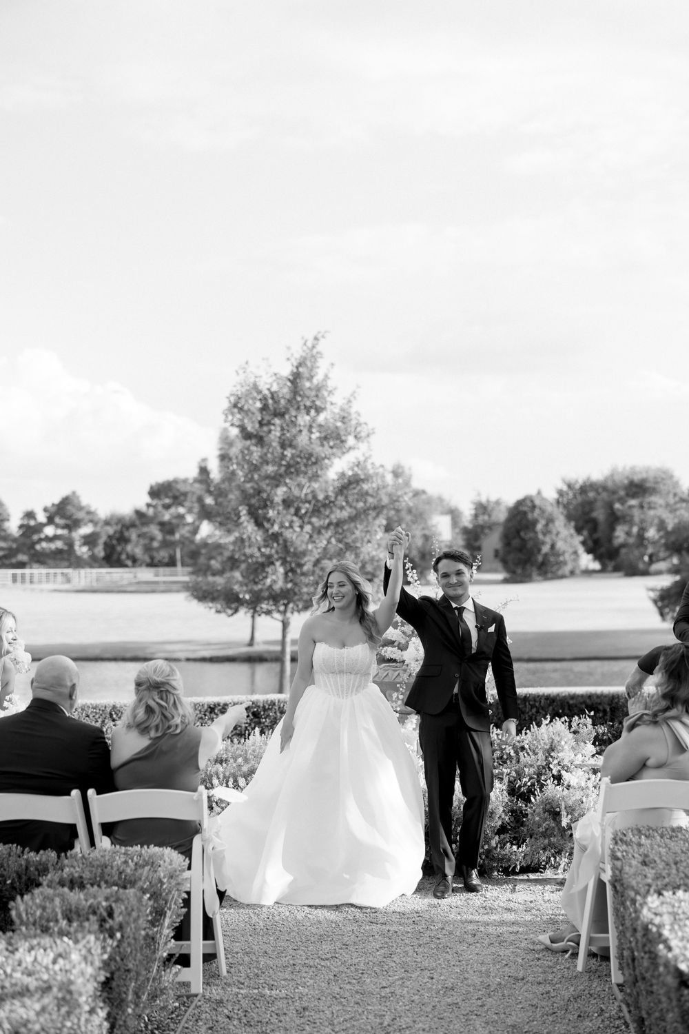 Wedding guests gather outdoors for a ceremony on a sunny day with white chairs arranged on manicured grounds.