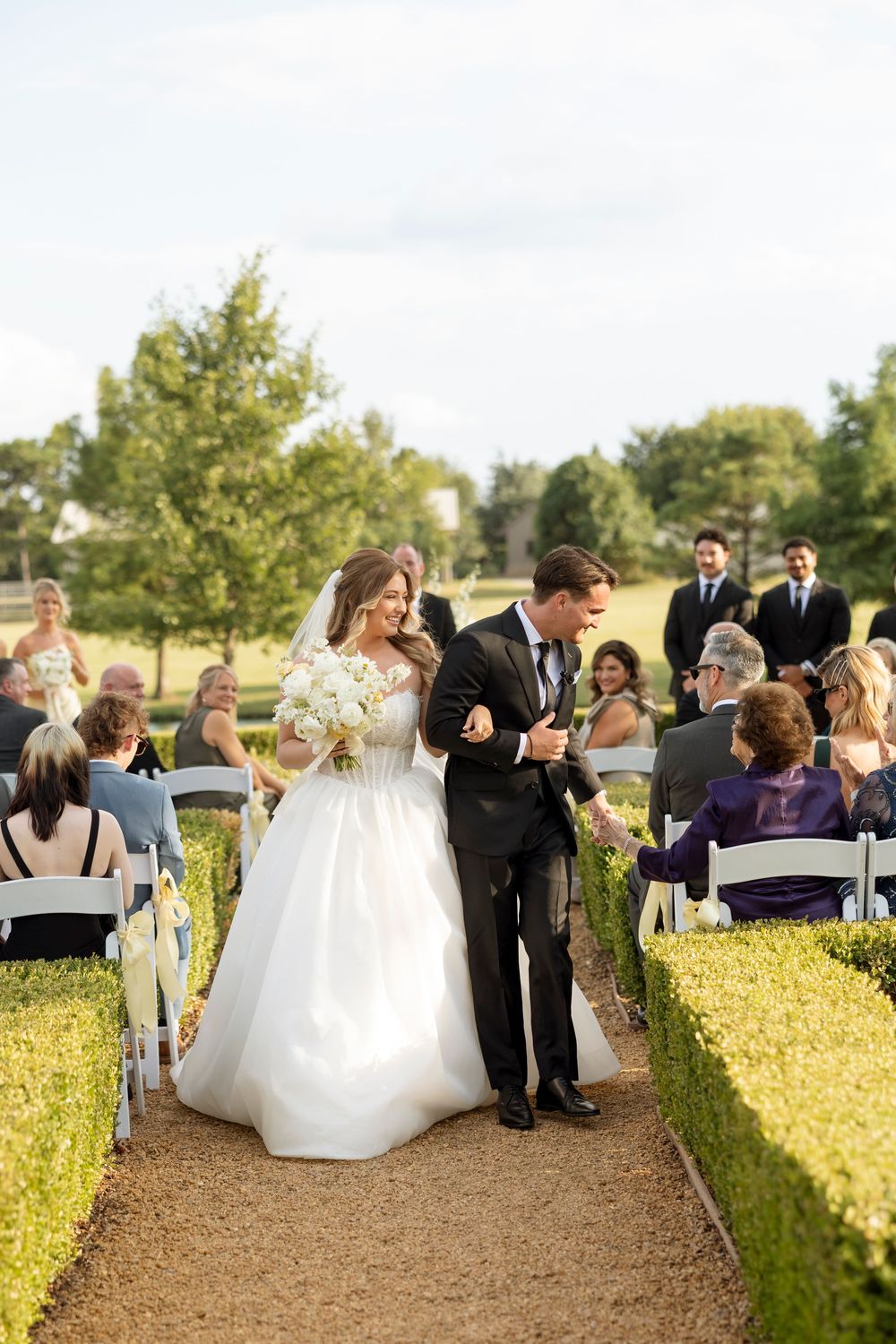 Wedding party and guests mingle on a garden path lined with yellow flowers and hedges during an outdoor celebration.