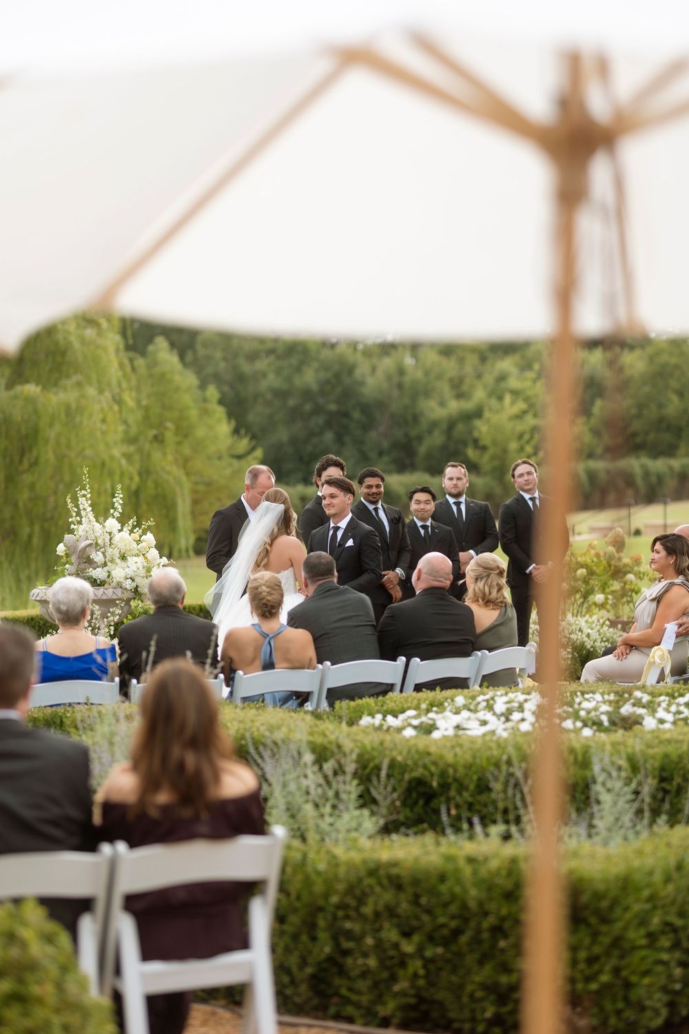 Outdoor wedding ceremony setup with white chairs arranged in rows surrounded by lush gardens and greenery.