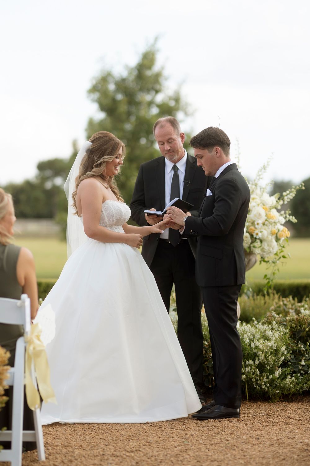 Outdoor wedding ceremony with the bridal party gathered on a gravel path surrounded by greenery and white chairs.