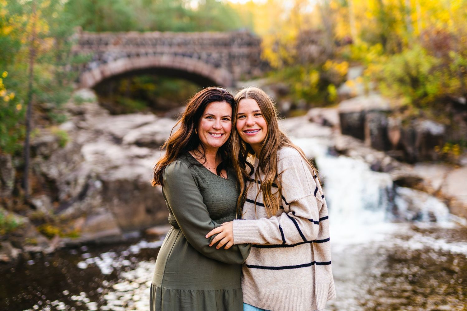 Two friends embrace and smile in front of a stone bridge and waterfall during autumn with golden foliage in background.