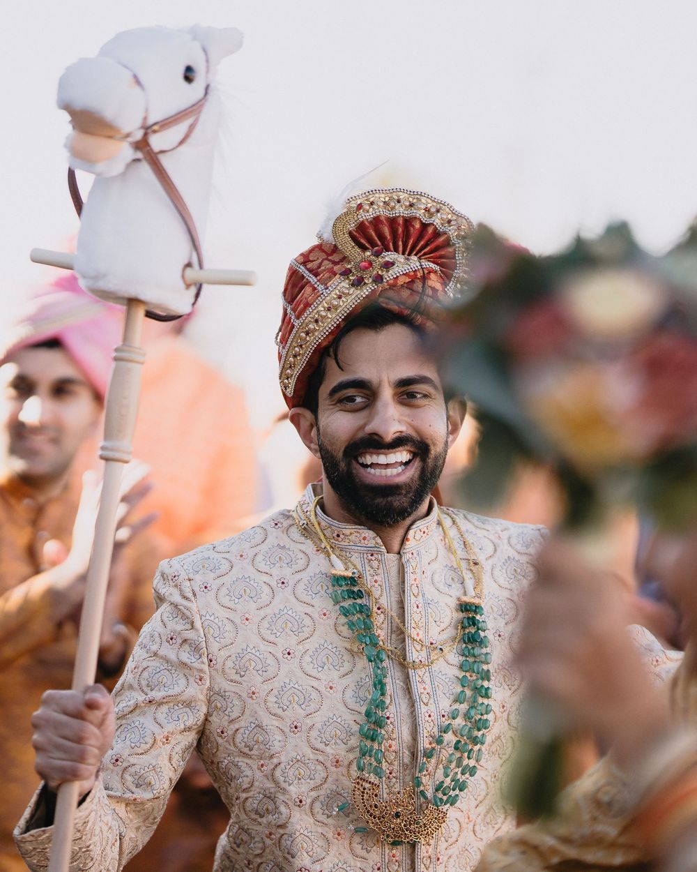 Smiling Indian groom in ivory sherwani holds hobby horse during traditional Baraat ceremony with emerald garland.at Sandia Resort and Casino