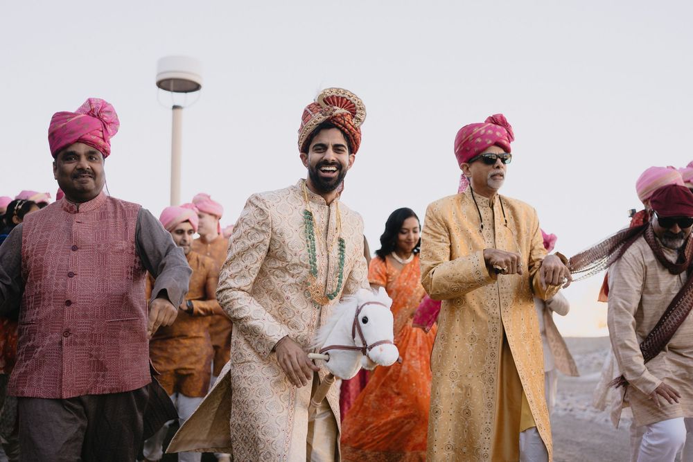 Indian groom in ivory sherwani carries hobby horse while walking with groomsmen in pink turbans at Baraat.at Sandia Resort and Casino