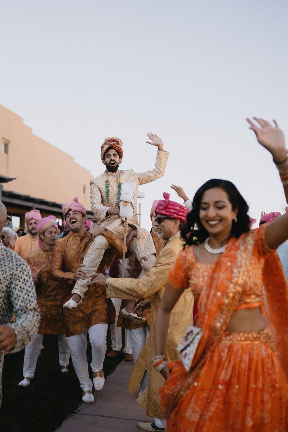 Indian groom lifted by groomsmen during joyful Baraat procession as woman in orange lehenga dances in foreground.at Sandia Resort and Casino