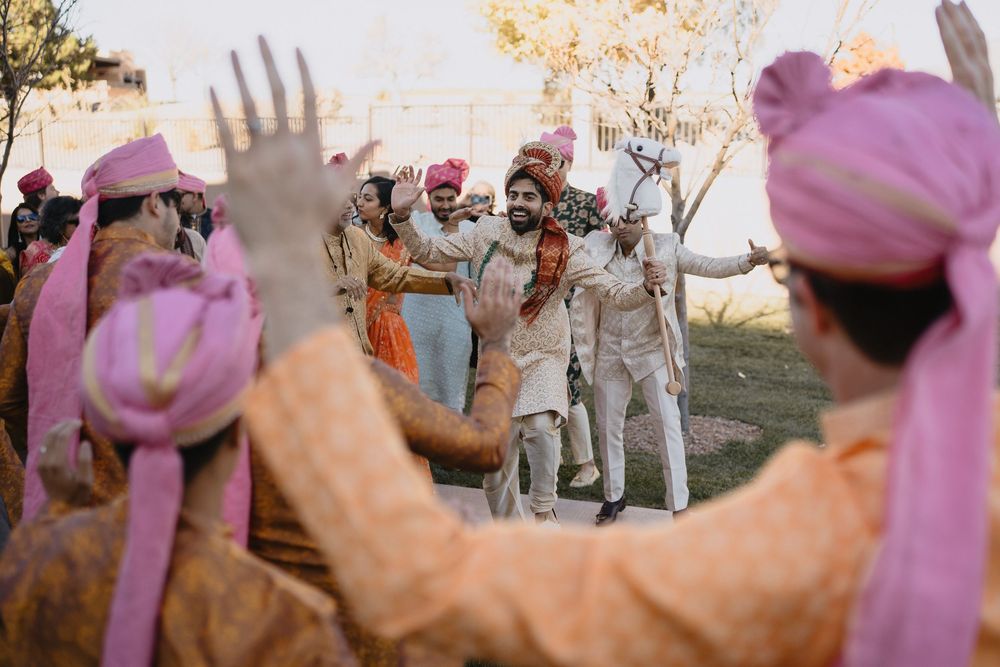 Indian groom in ivory sherwani arrives at Baraat ceremony as guests in orange kurtas raise hands in celebration.at Sandia Resort and Casino