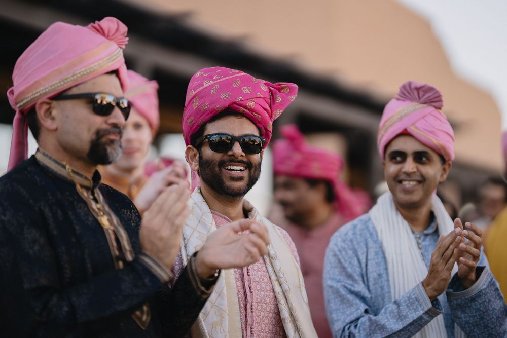 Three men in pink turbans and sunglasses clap and smile joyfully during an Indian wedding Baraat celebration.