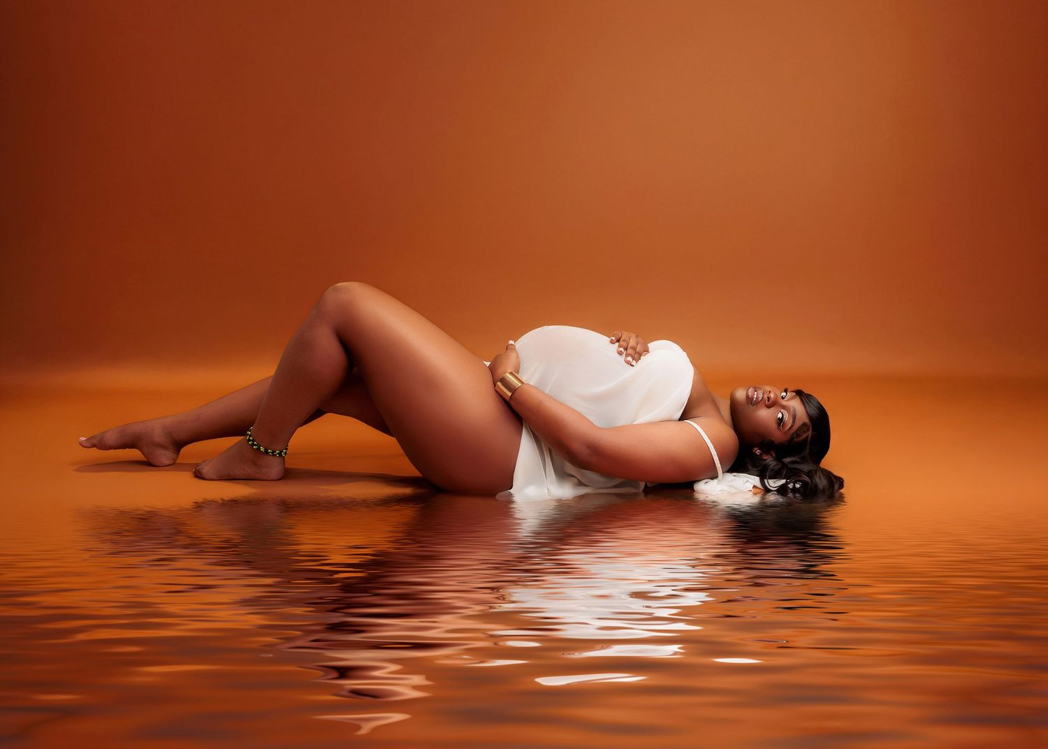 Pregnant woman in white draped fabric lying gracefully on reflective orange studio floor.