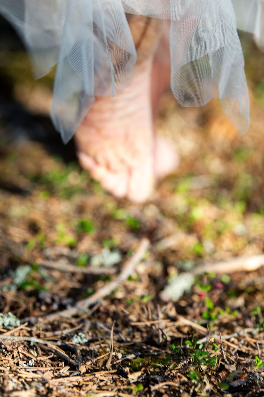 Close-up view of bare feet and white tulle fabric against forest ground.