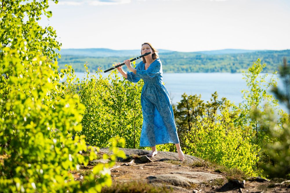 A flutist performs on a scenic overlook with mountains and water in the background.