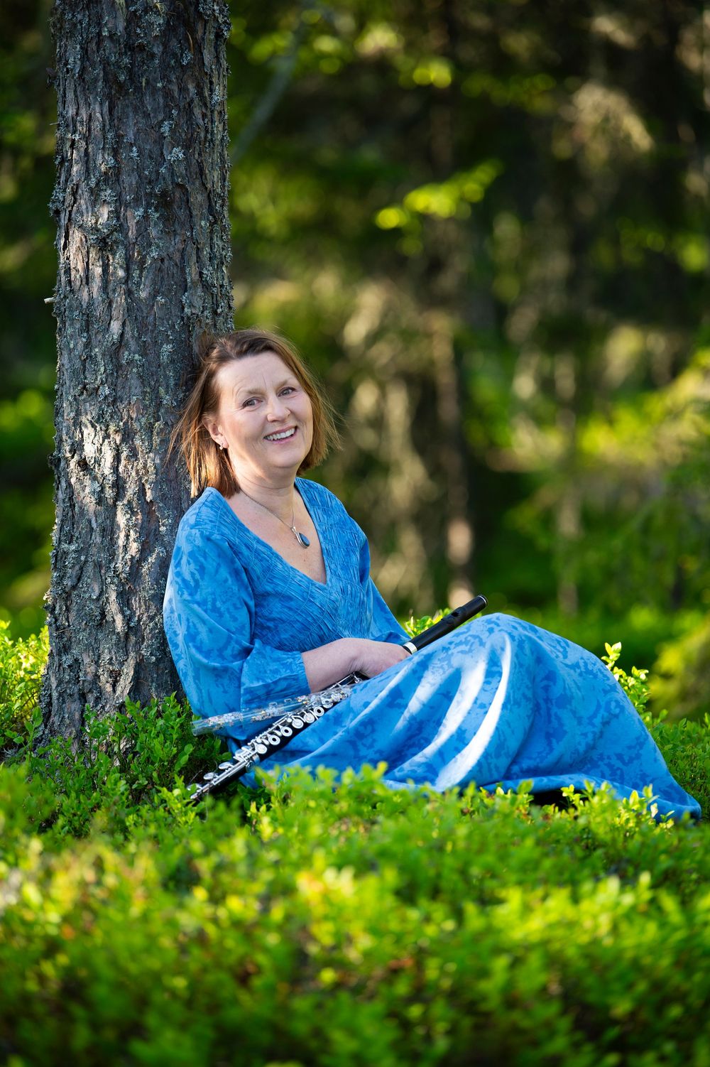 Someone in a bright blue dress reclines against a tree trunk in dappled sunlight.
