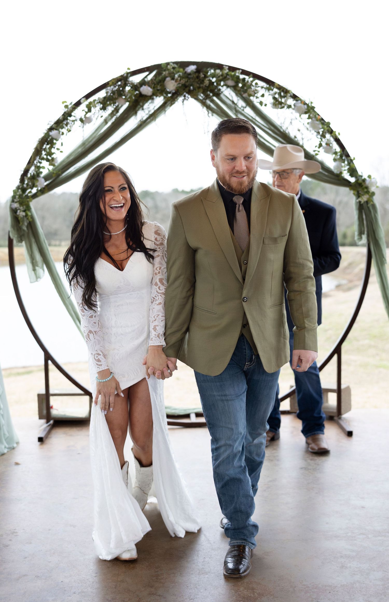 A couple walks together under a circular wedding arch decorated with greenery during their rustic ceremony.