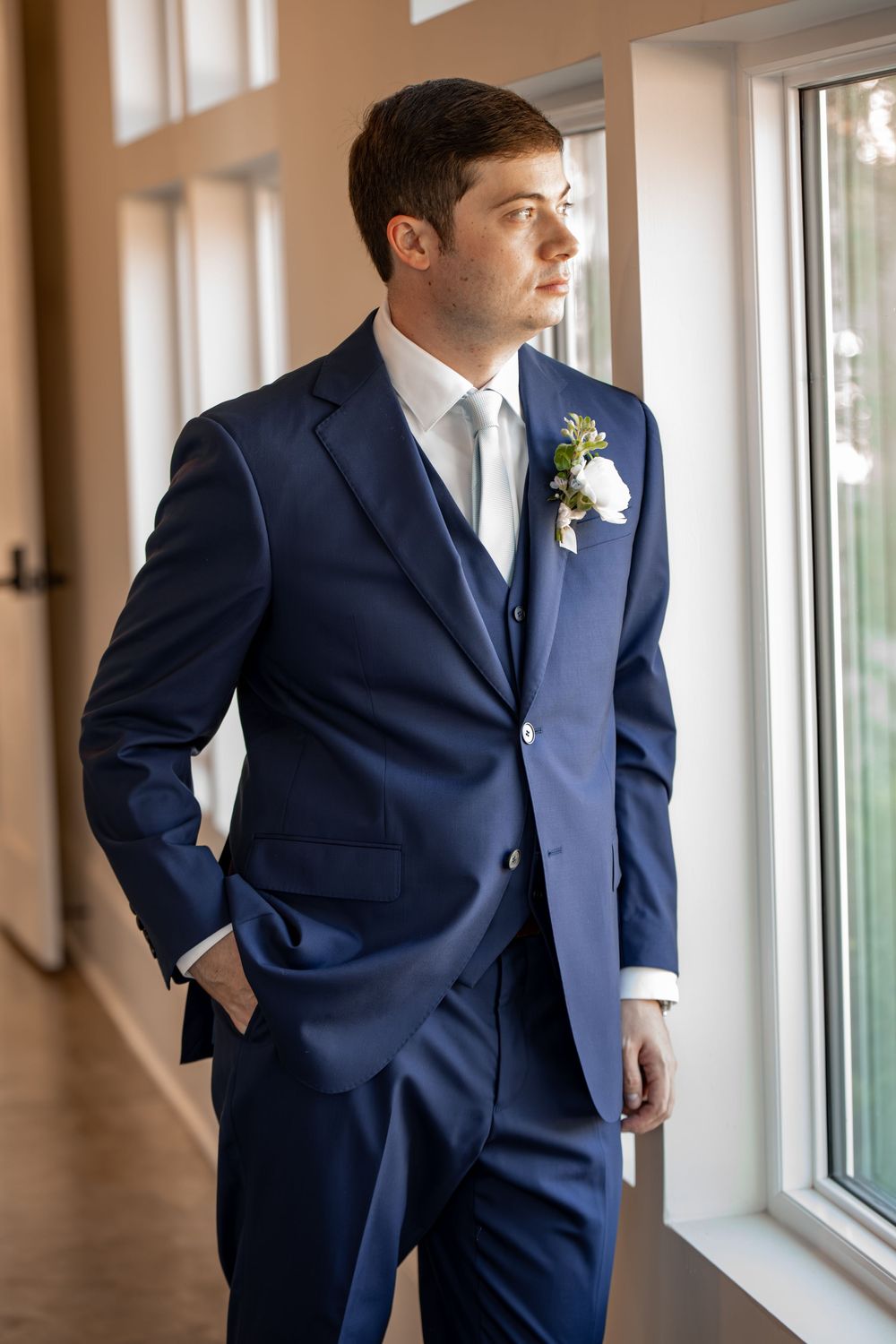 A groom in a navy blue suit stands thoughtfully by a window in an elegant hallway.