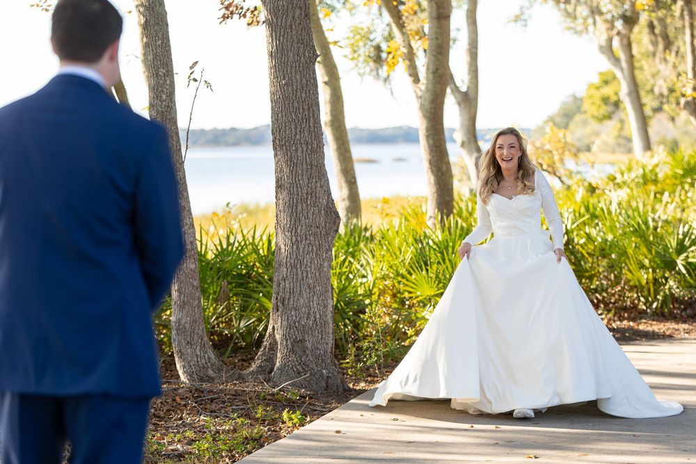 Bride in flowing white gown walks along waterfront path lined with trees.