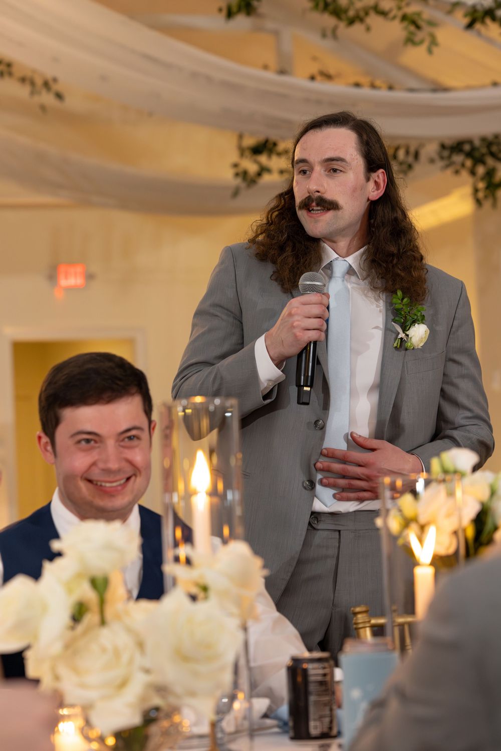 Wedding party members in gray suits and navy suits give toasts during an indoor reception celebration.