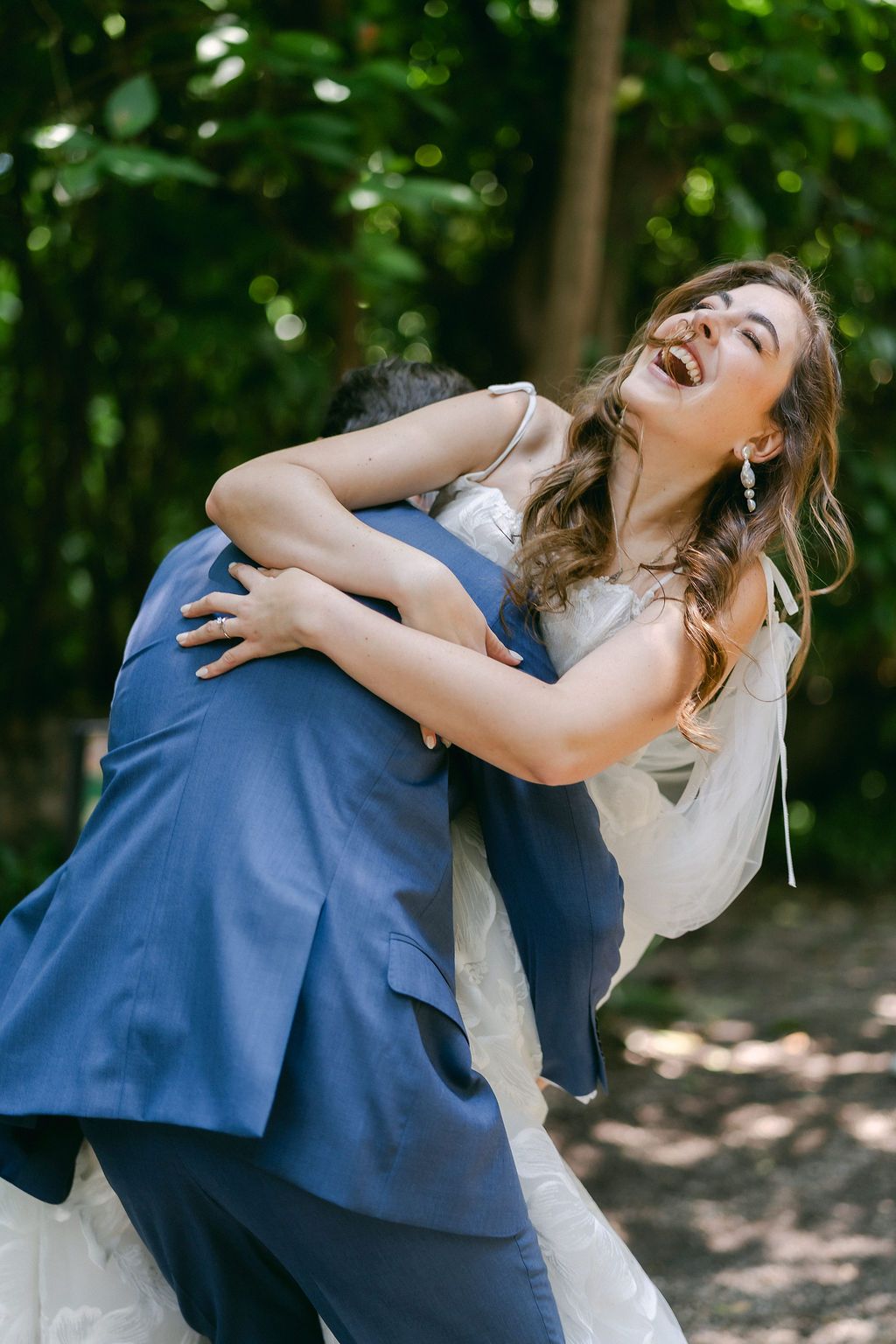 A couple embraces joyfully outdoors in wedding attire, with the bride in white and groom in navy blue against a forest backdrop.