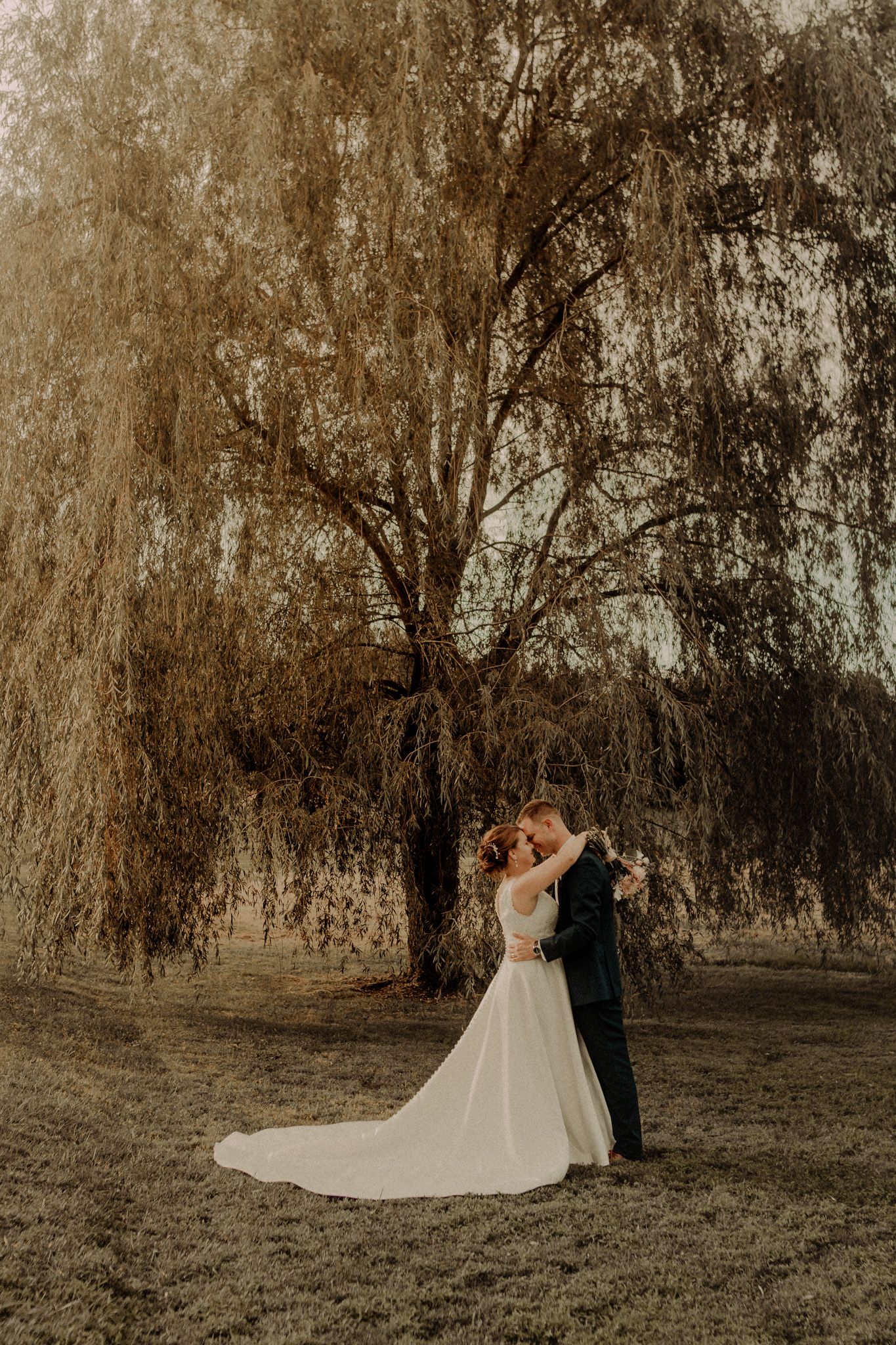 A romantic wedding couple stands beneath drooping willow trees at sunset creating a dreamy outdoor portrait.