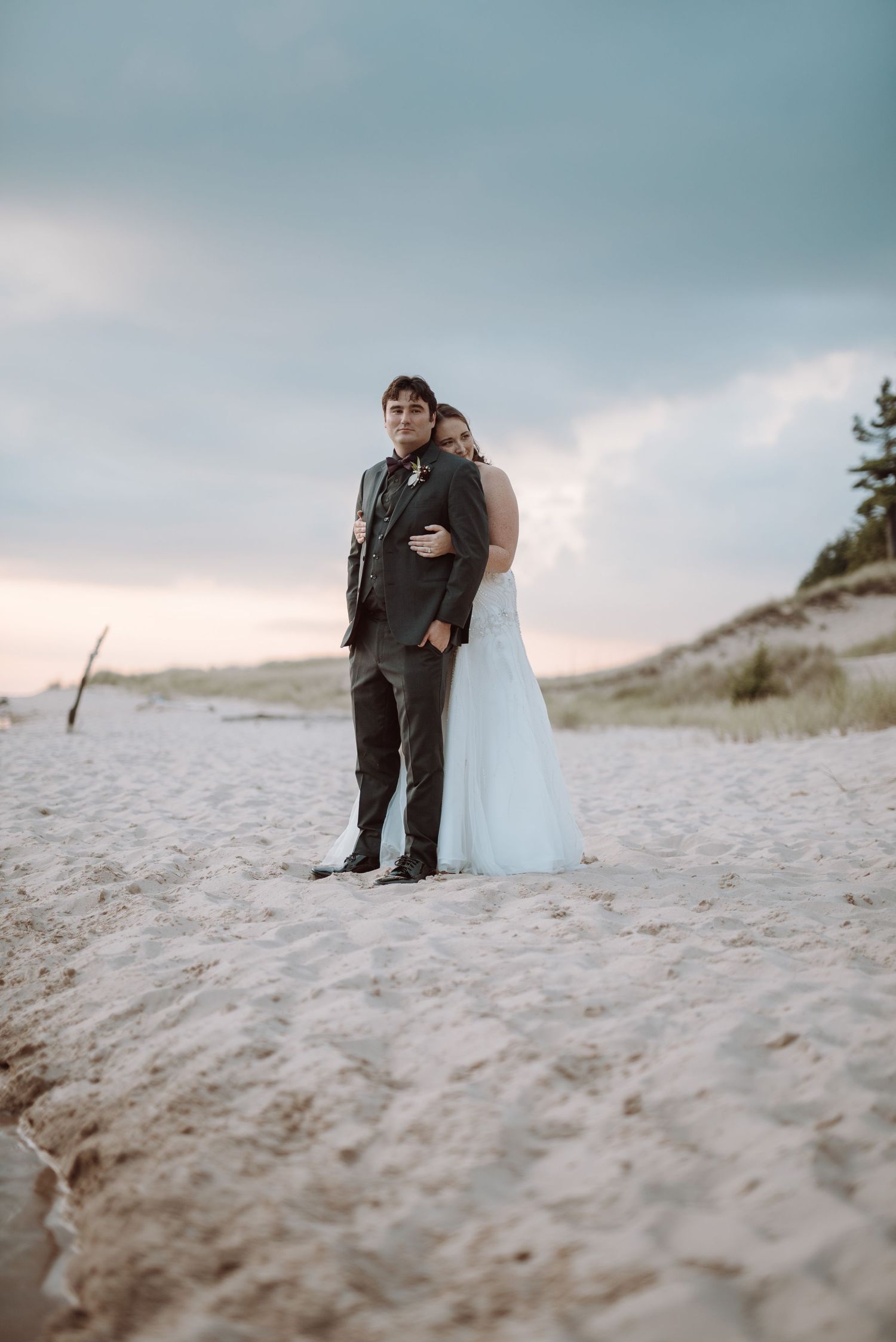 A couple embraces on a sandy beach during their winter wedding photoshoot against a moody cloudy sky.