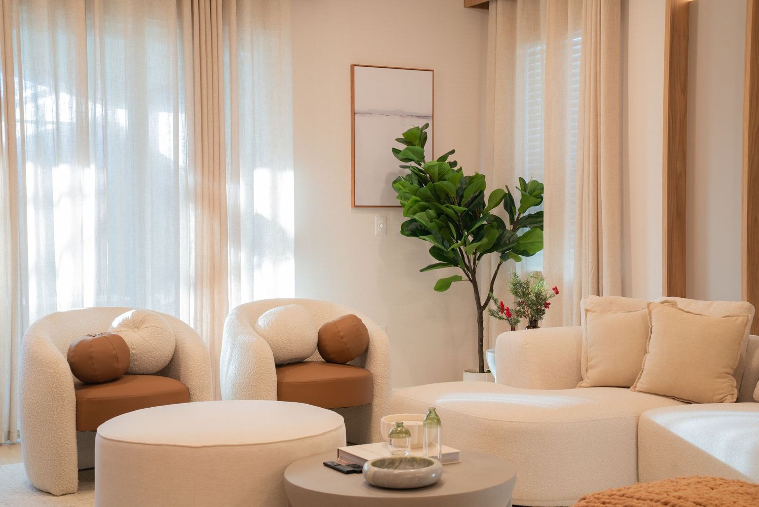 Modern living room interior with beige furniture, fiddle leaf fig plant, and natural light streaming through sheer curtains.
