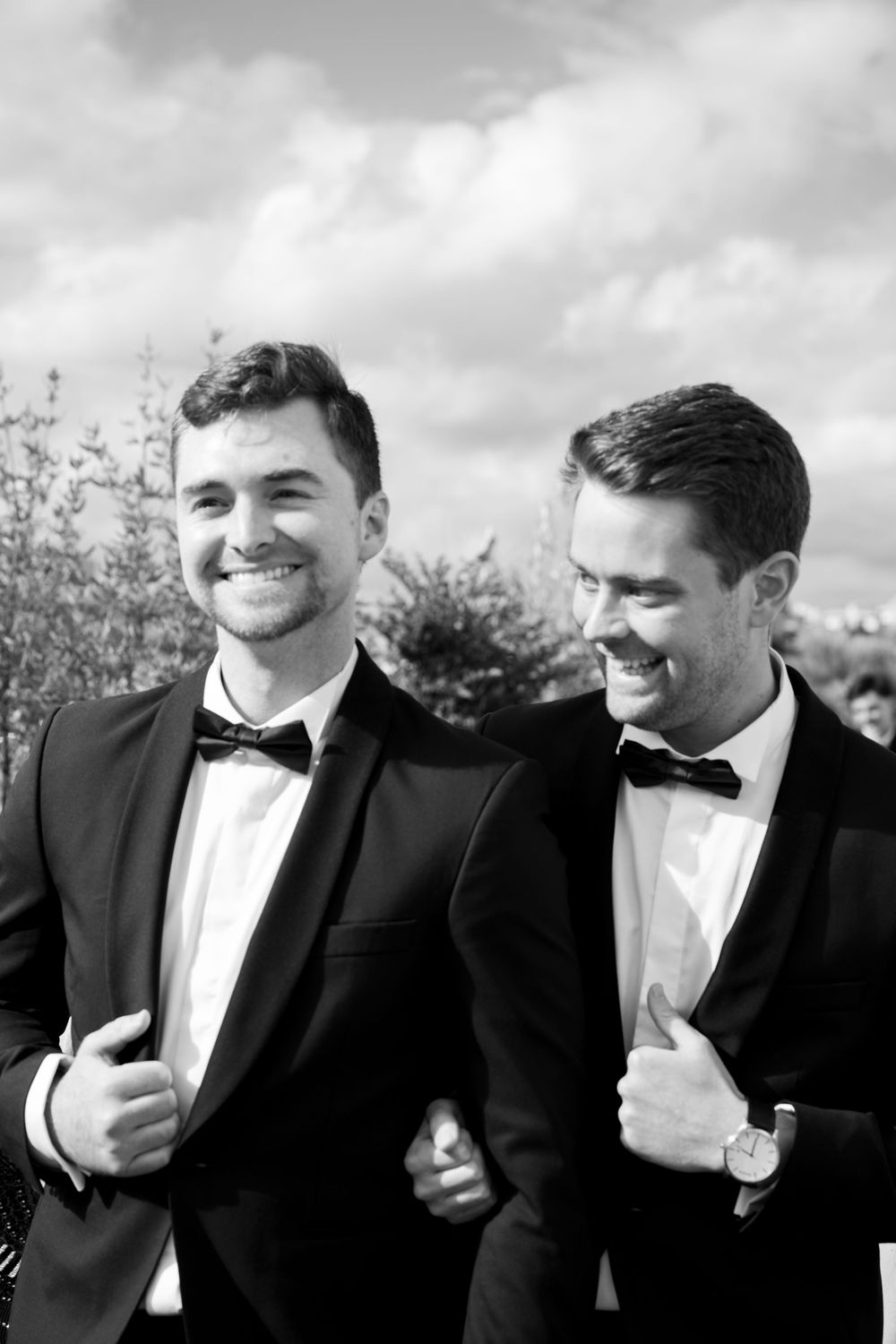 Groomsmen in black tuxedos and bow ties share a candid laughing moment together in black and white photography.