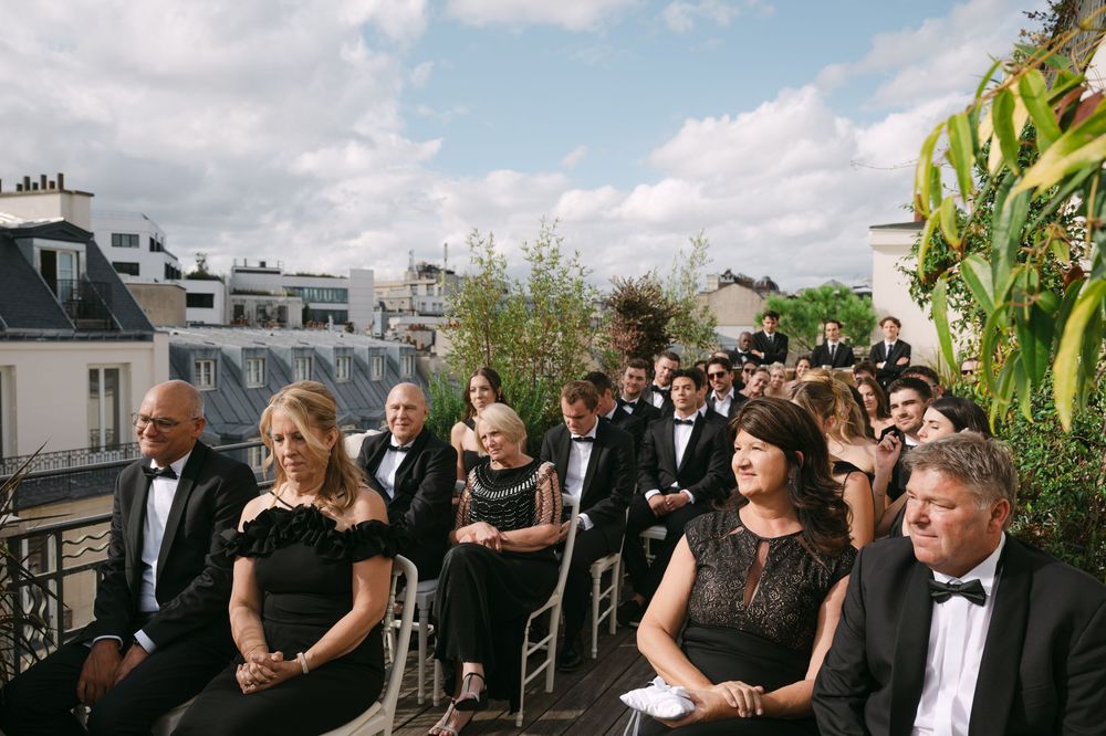 A panoramic view of wedding guests seated outdoors at a garden ceremony with greenery and buildings in the background.