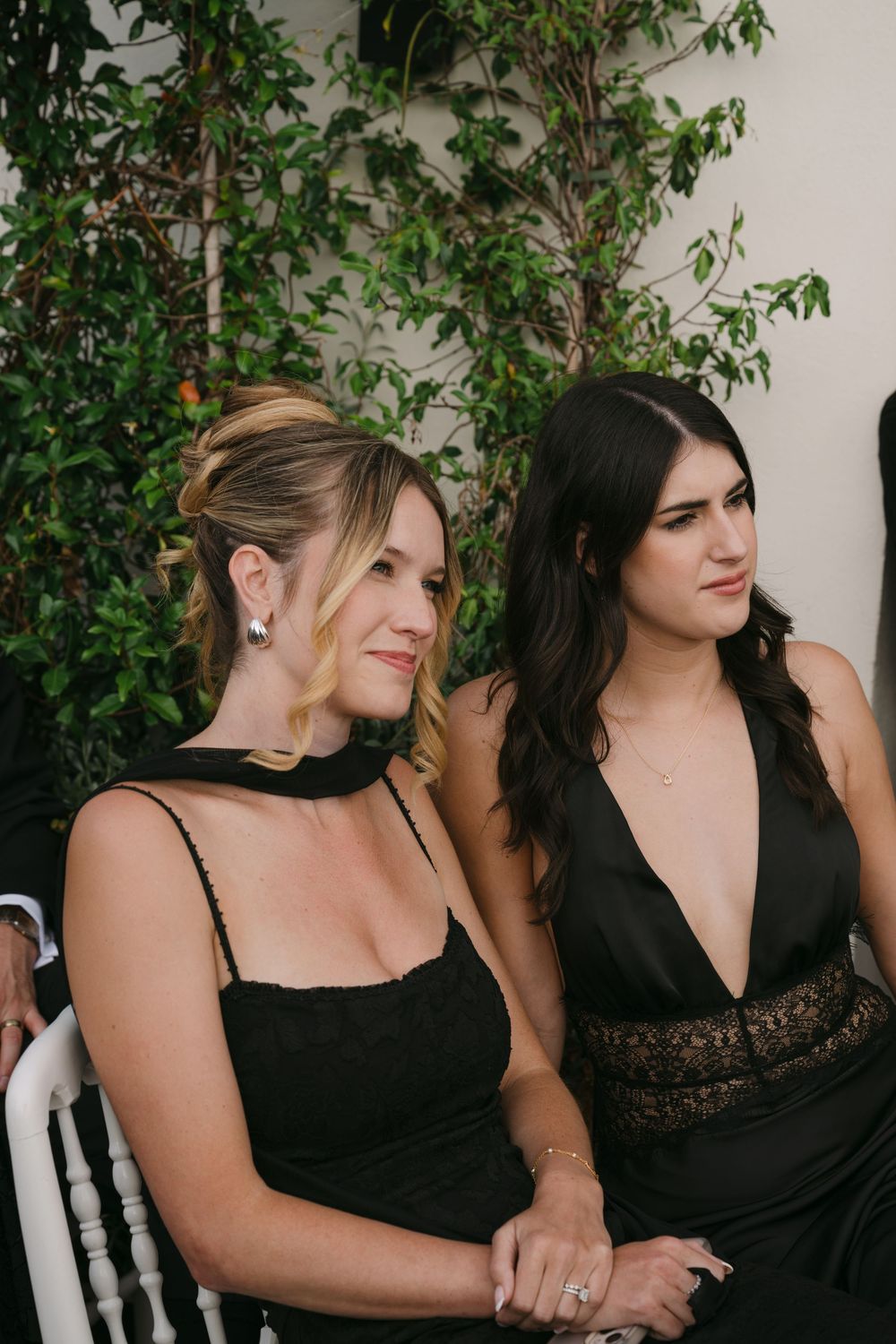 A group of women in black dresses stand together against a white wall with green foliage in the background.