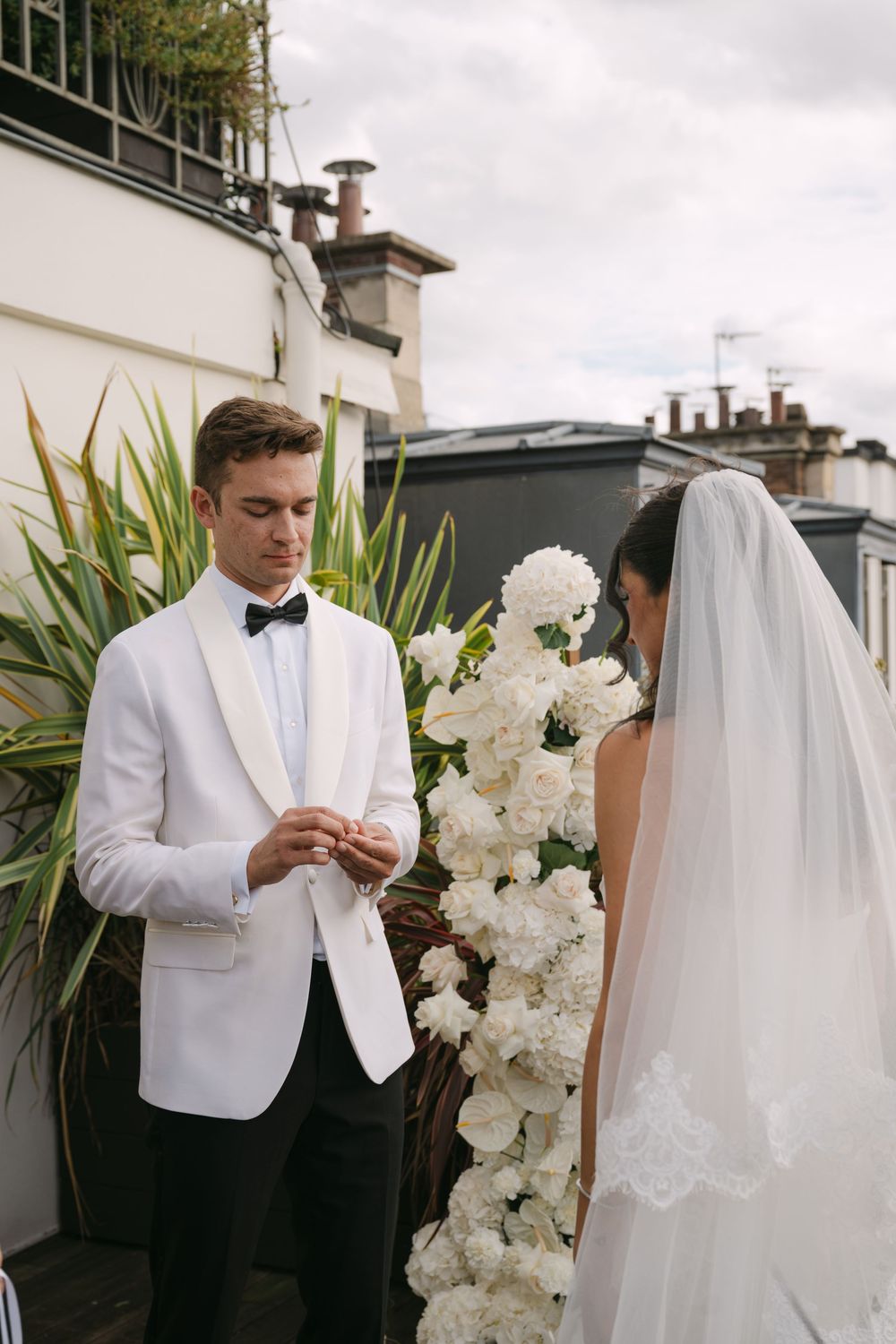 A wedding couple exchanges rings during an outdoor ceremony surrounded by white floral arrangements.