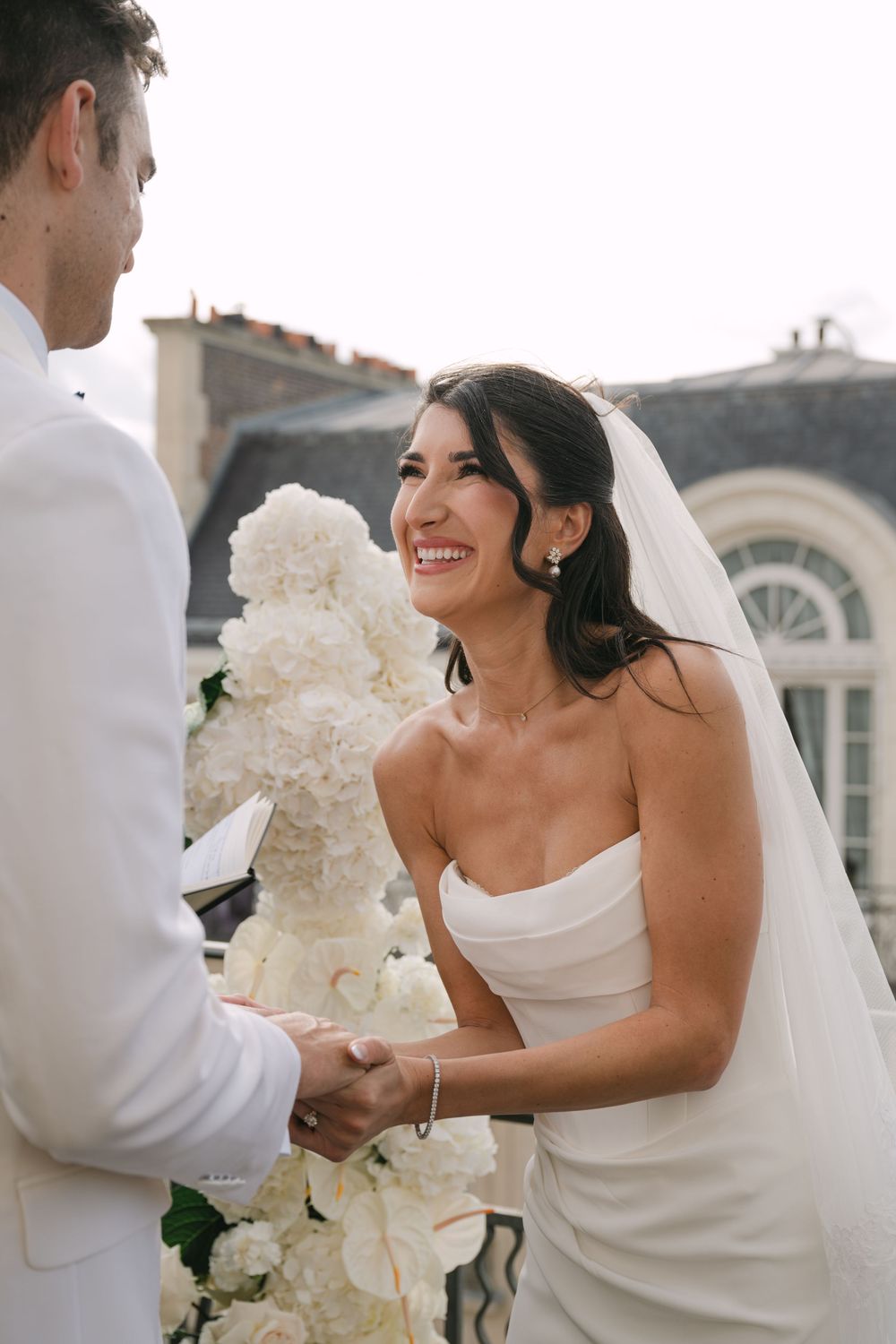 A joyful wedding moment captured as the bride in a white gown holds a bouquet of white flowers during an outdoor ceremony.