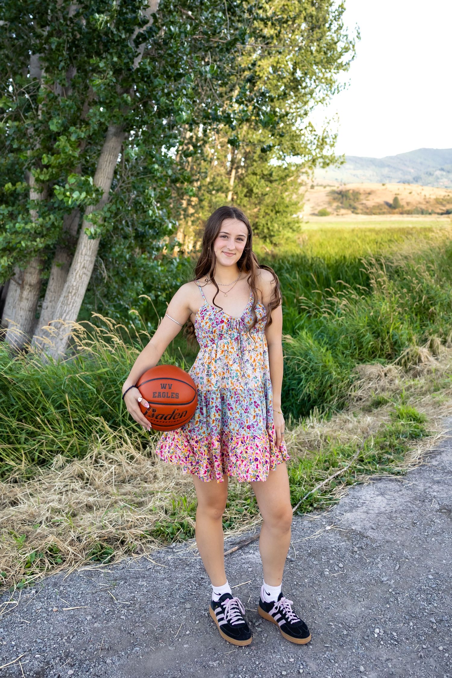 A basketball player in a floral sundress poses with a basketball on a path near trees and mountains.