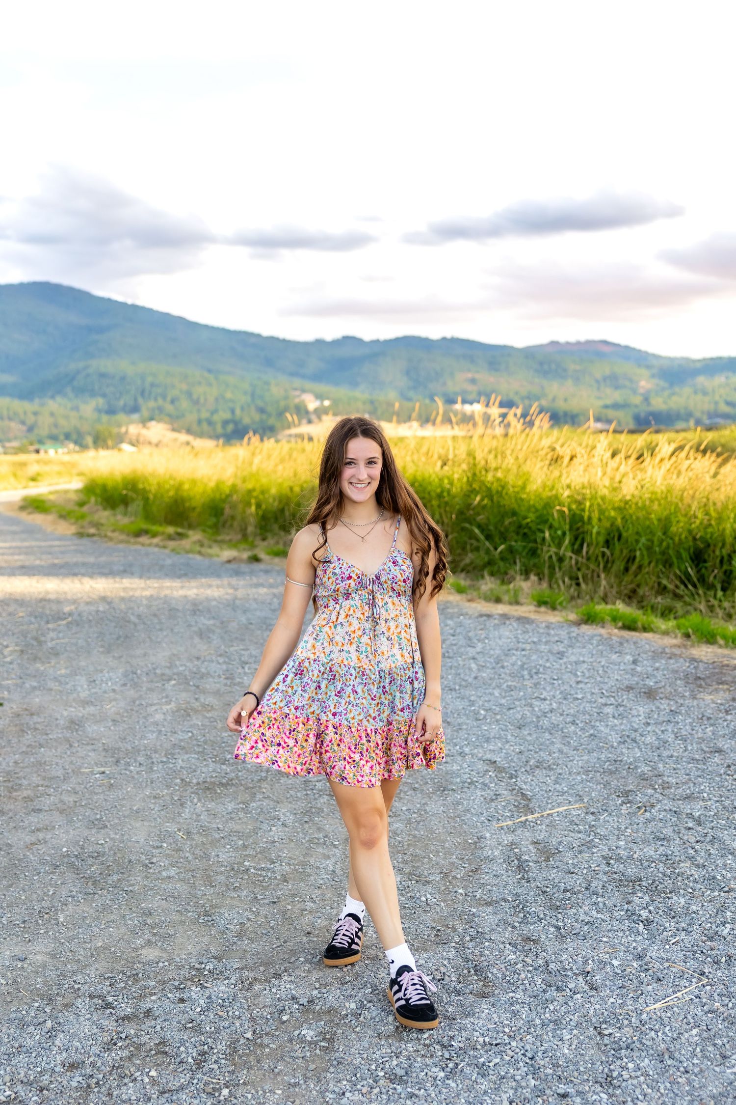 A person in a floral sundress and sneakers stands on a gravel path with mountains and golden grass in the background.