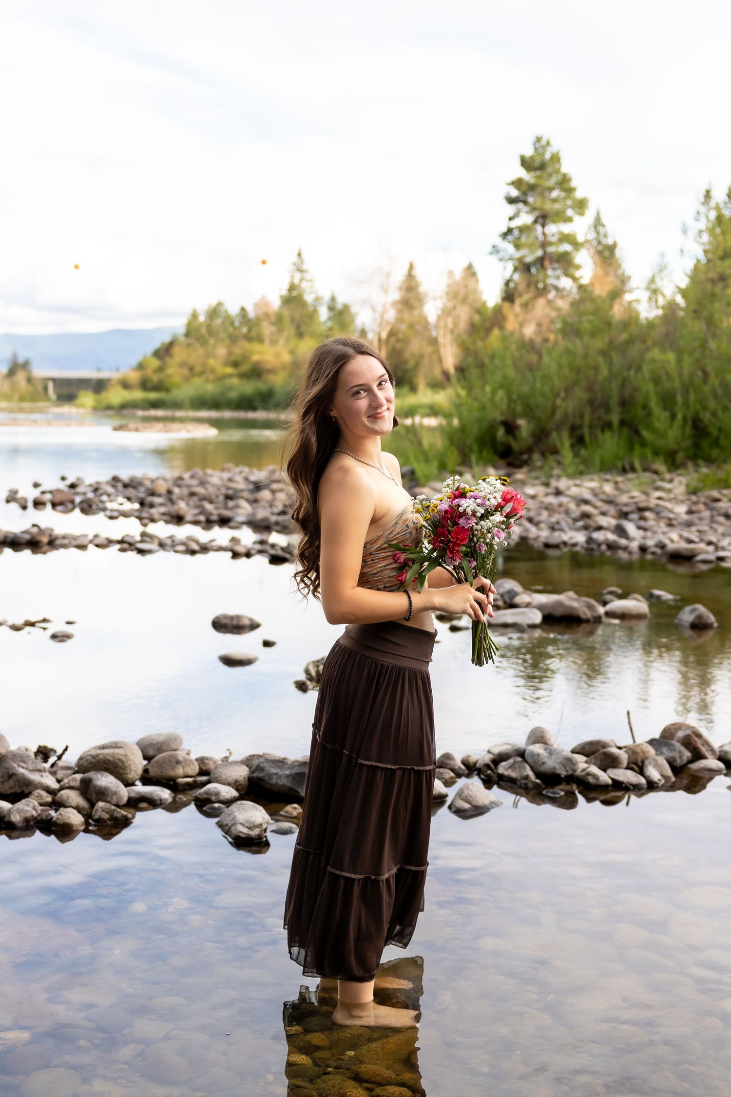 A figure in a brown dress poses with flowers by a rocky lakeshore with evergreen trees in the background.
