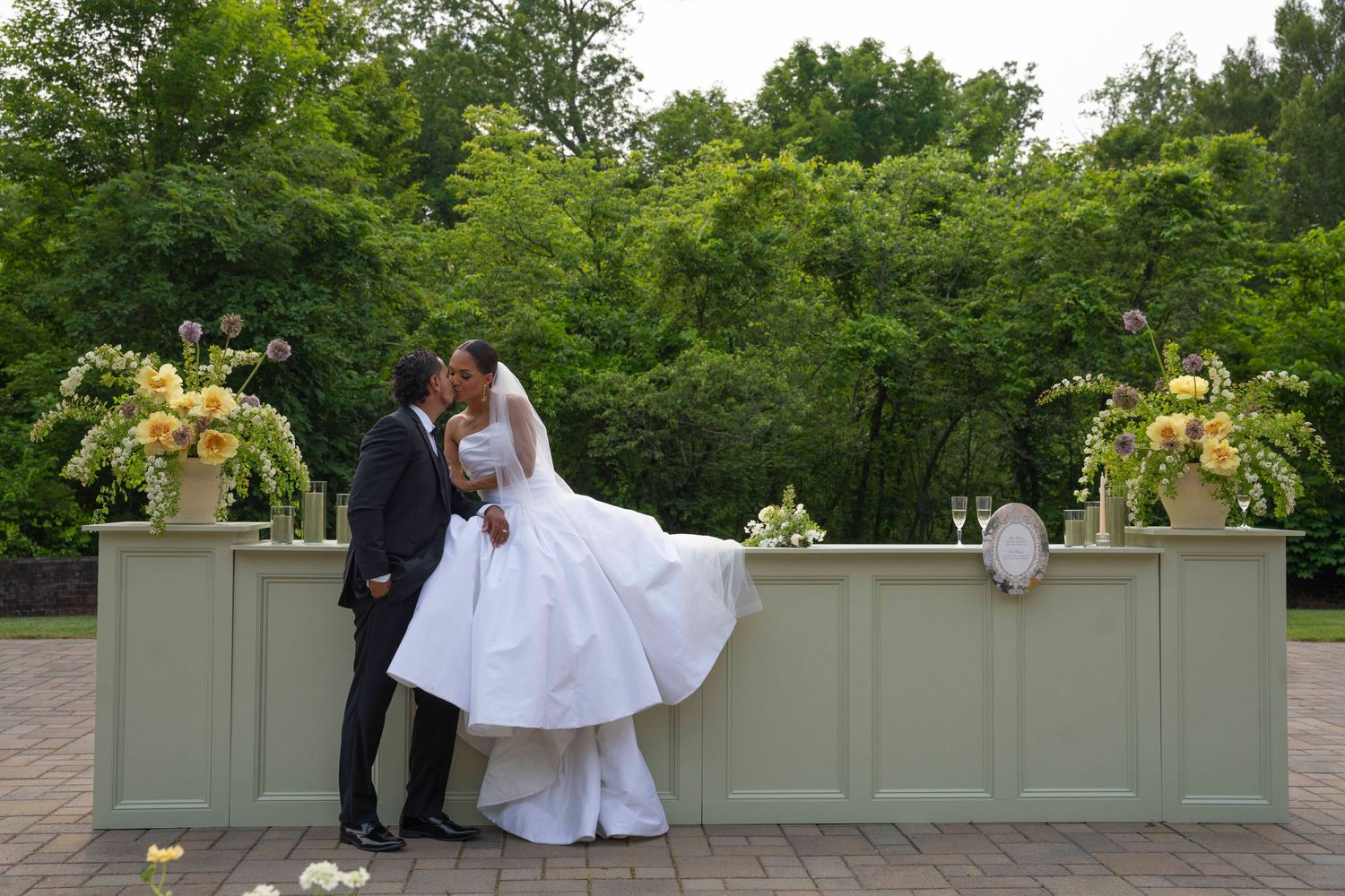 A romantic outdoor wedding scene with a couple in formal attire sharing a tender moment on a stone terrace.