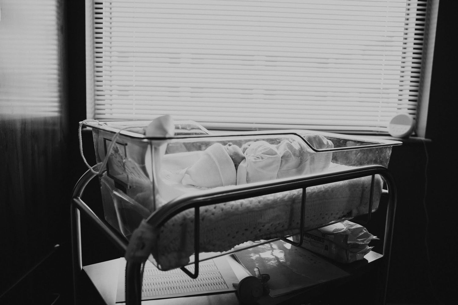 Black and white photo of a newborn baby sleeping peacefully in a hospital bassinet near a window.