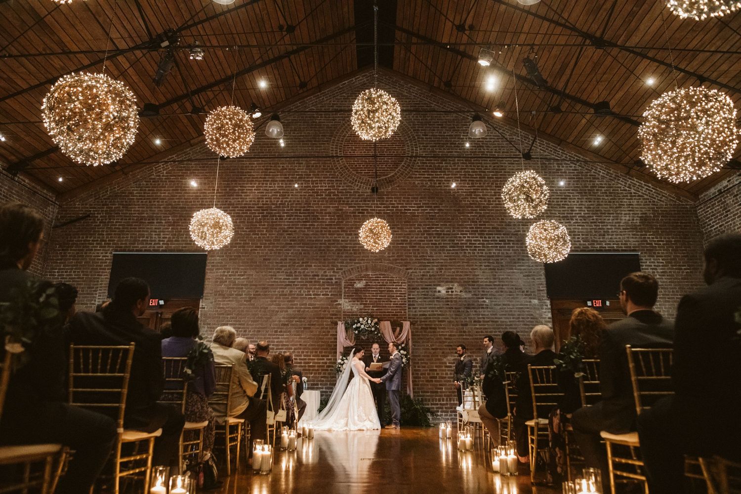 Rustic brick venue decorated with illuminated sphere chandeliers and white table settings for an evening event.