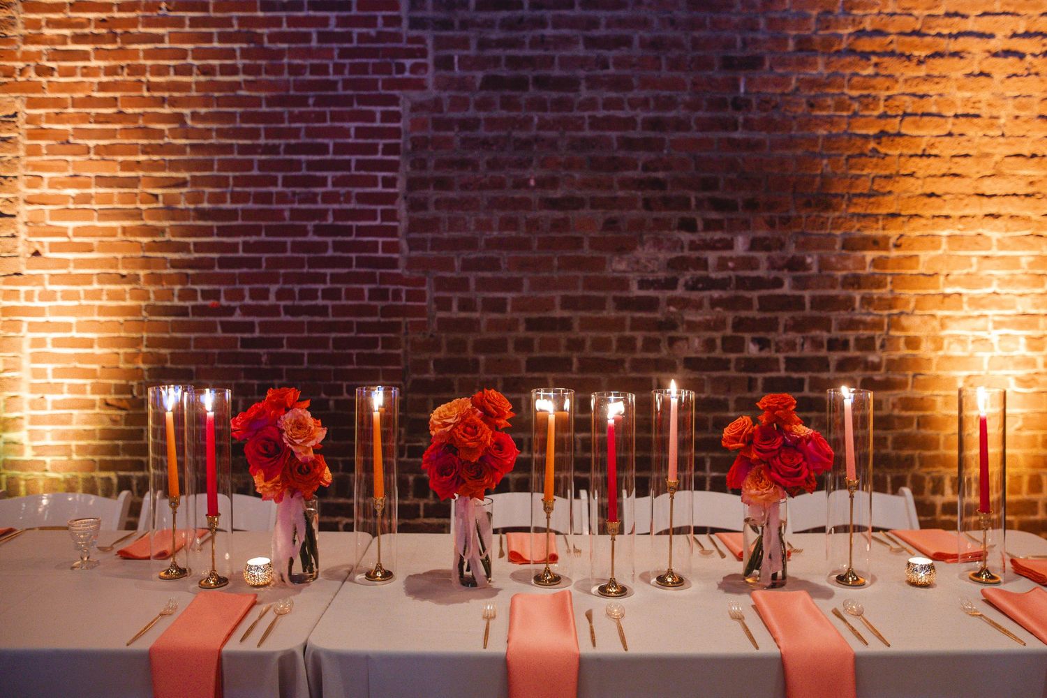 Elegant wedding reception table setup with red floral centerpieces against exposed brick wall illuminated by warm lighting.
