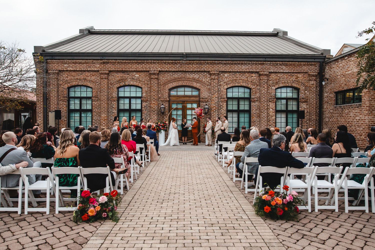 Wedding ceremony setup with white chairs and floral decorations outside a historic brick building with arched windows.