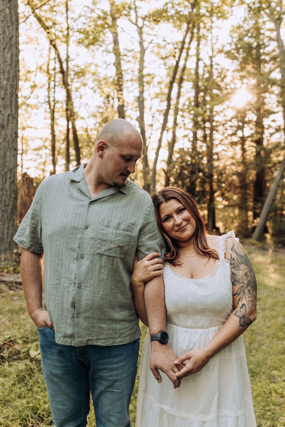 Couple posing in sunlit forest, woman in white dress with tattoos leans on mans shoulder smiling.