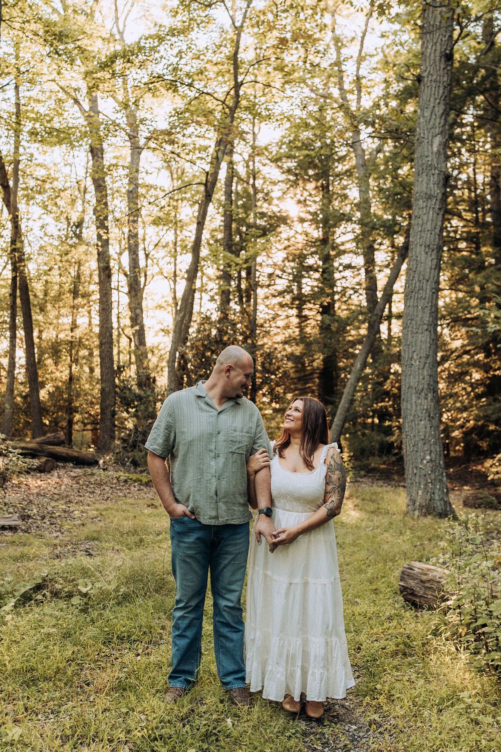 Engaged couple standing together in golden hour forest light, woman in white dress with floral tattoos.