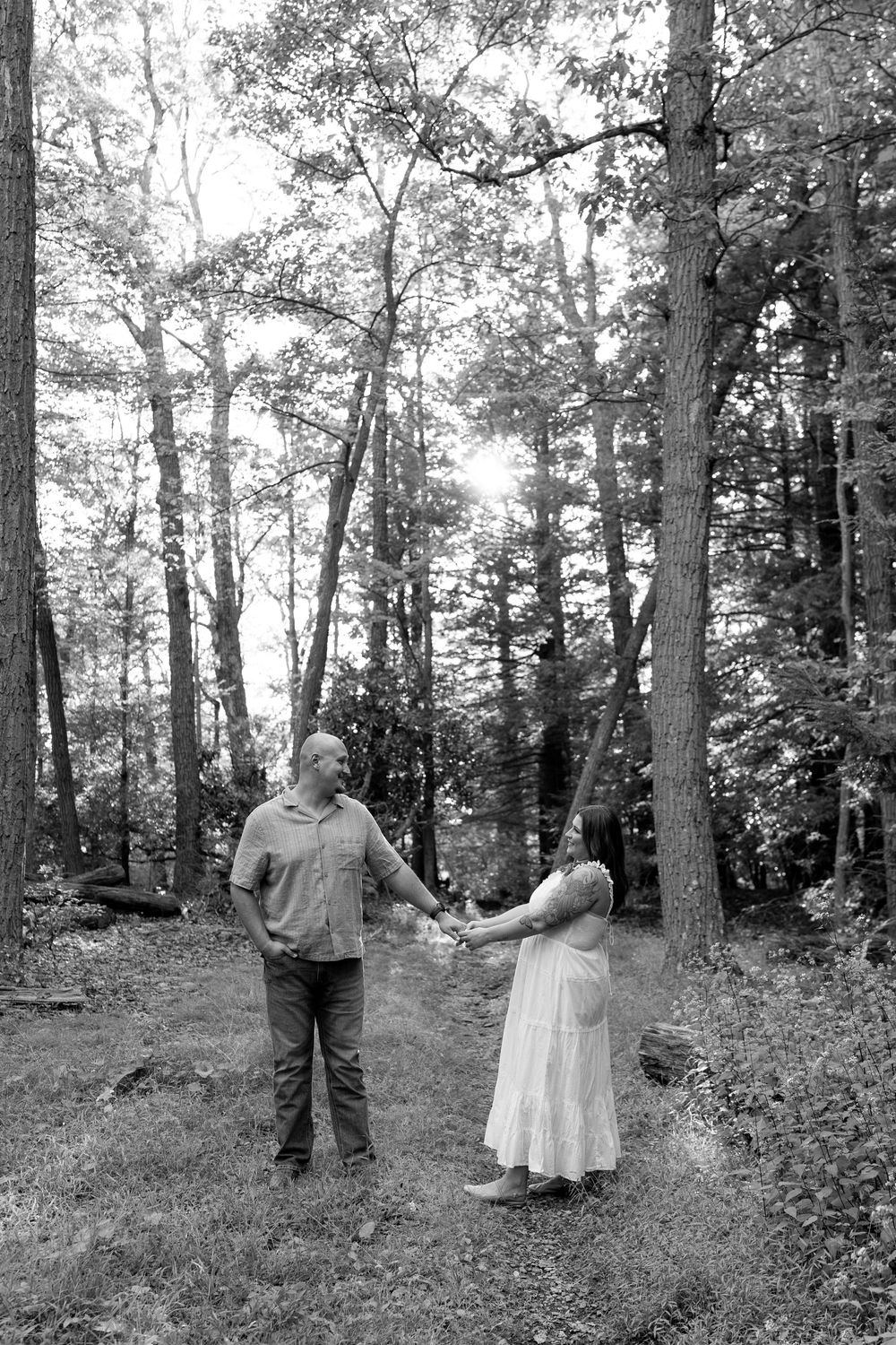 Black and white photo of couple holding hands in a wooded forest clearing, smiling at each other.