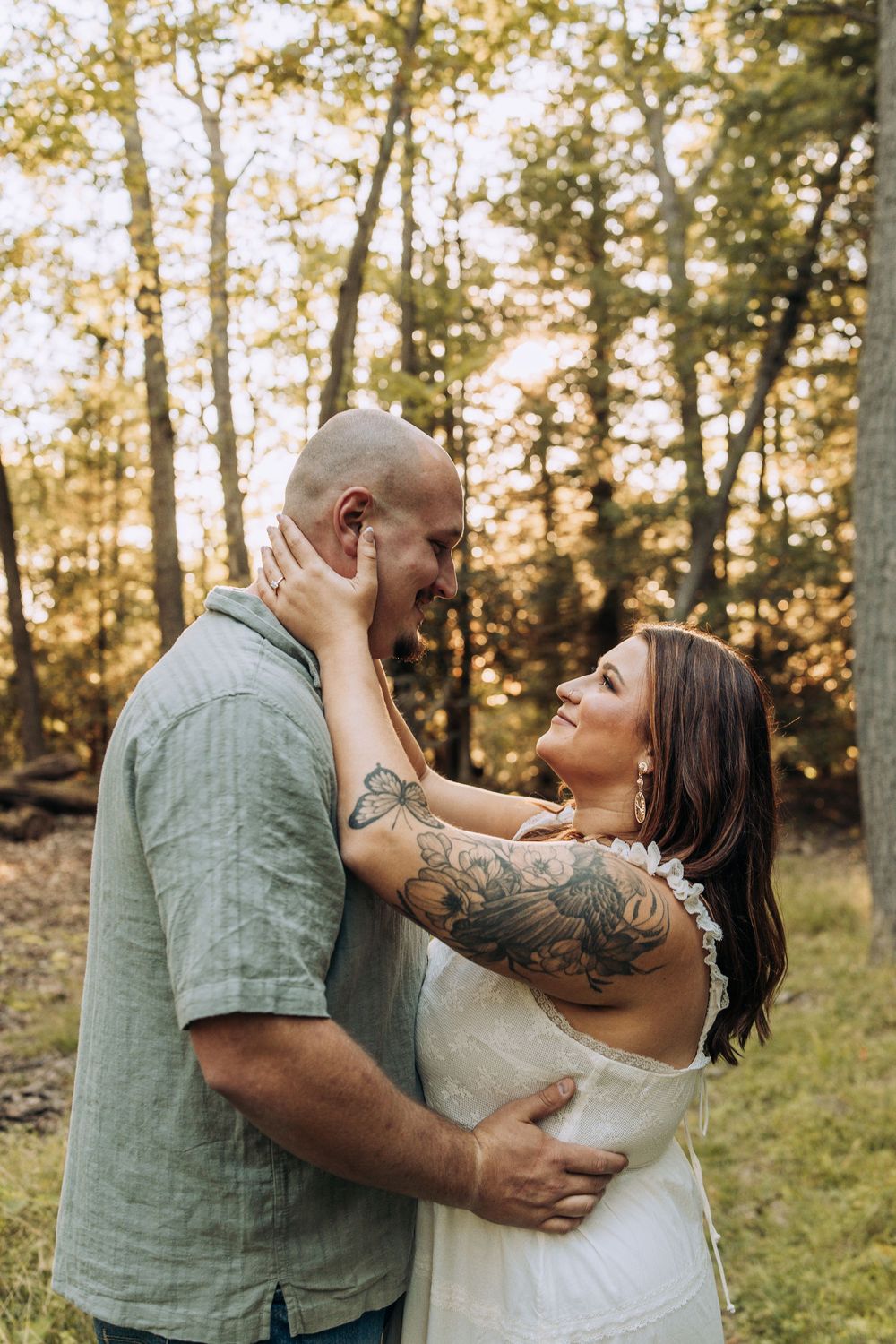 Laughing couple embracing in sun-dappled forest, tattooed woman in white dress holds mans face joyfully.