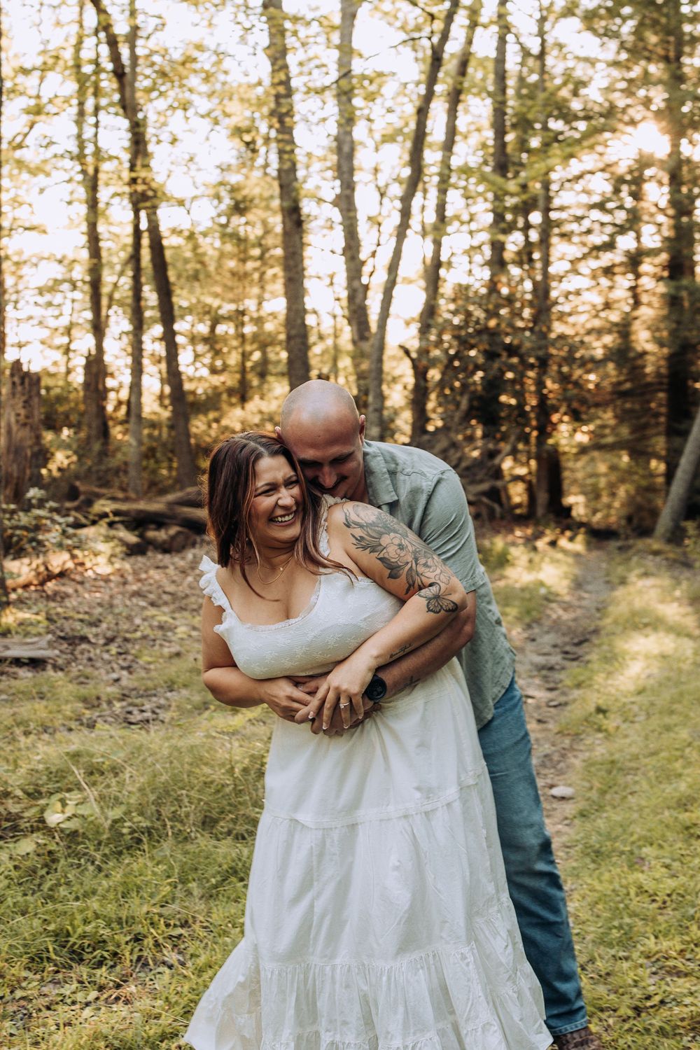 Joyful couple in forest at golden hour, man hugs woman in white dress from behind as she laughs.