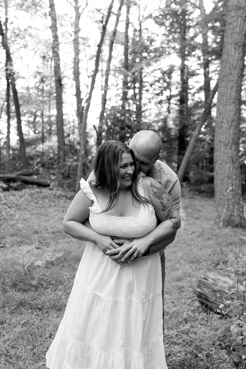 Black and white photo of couple embracing in woods, man kissing womans cheek as she smiles warmly.