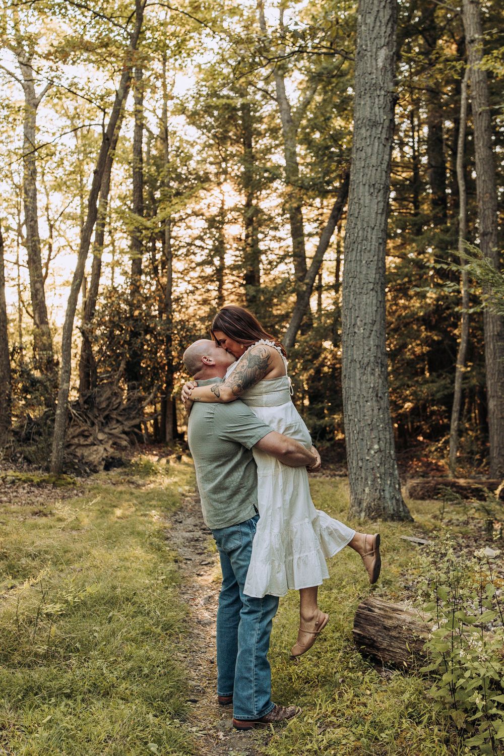 Man lifts woman in white dress for a kiss on a sunlit forest trail during golden hour.