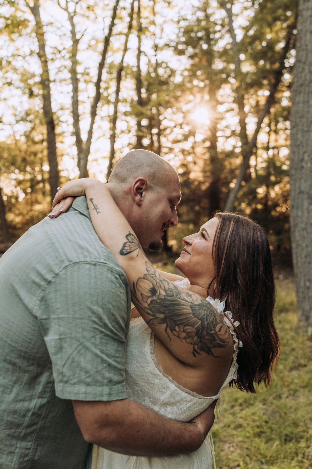 Couple sharing a romantic nose-to-nose moment in glowing sunset forest light, woman with floral arm tattoo.