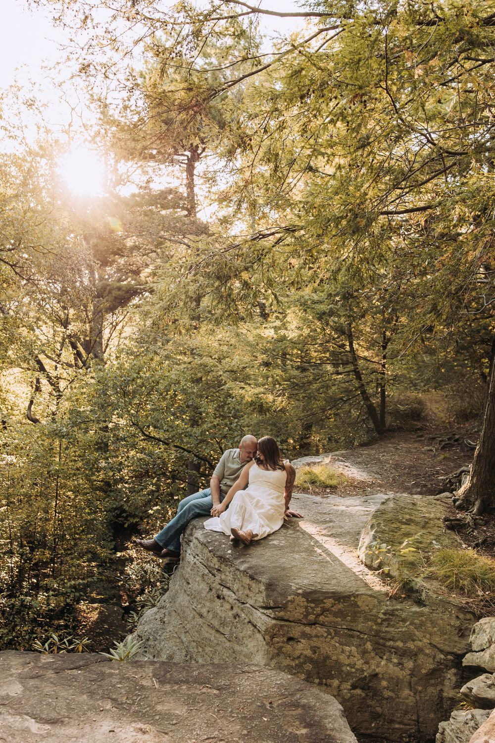 Couple sitting on mossy rocks kissing in a lush sunlit forest with golden rays filtering through trees.