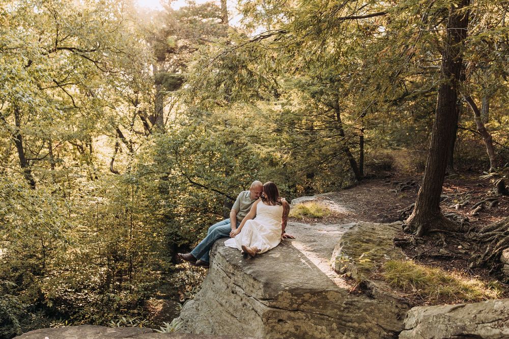 Couple sitting on a rock ledge surrounded by lush green trees in a forest setting.