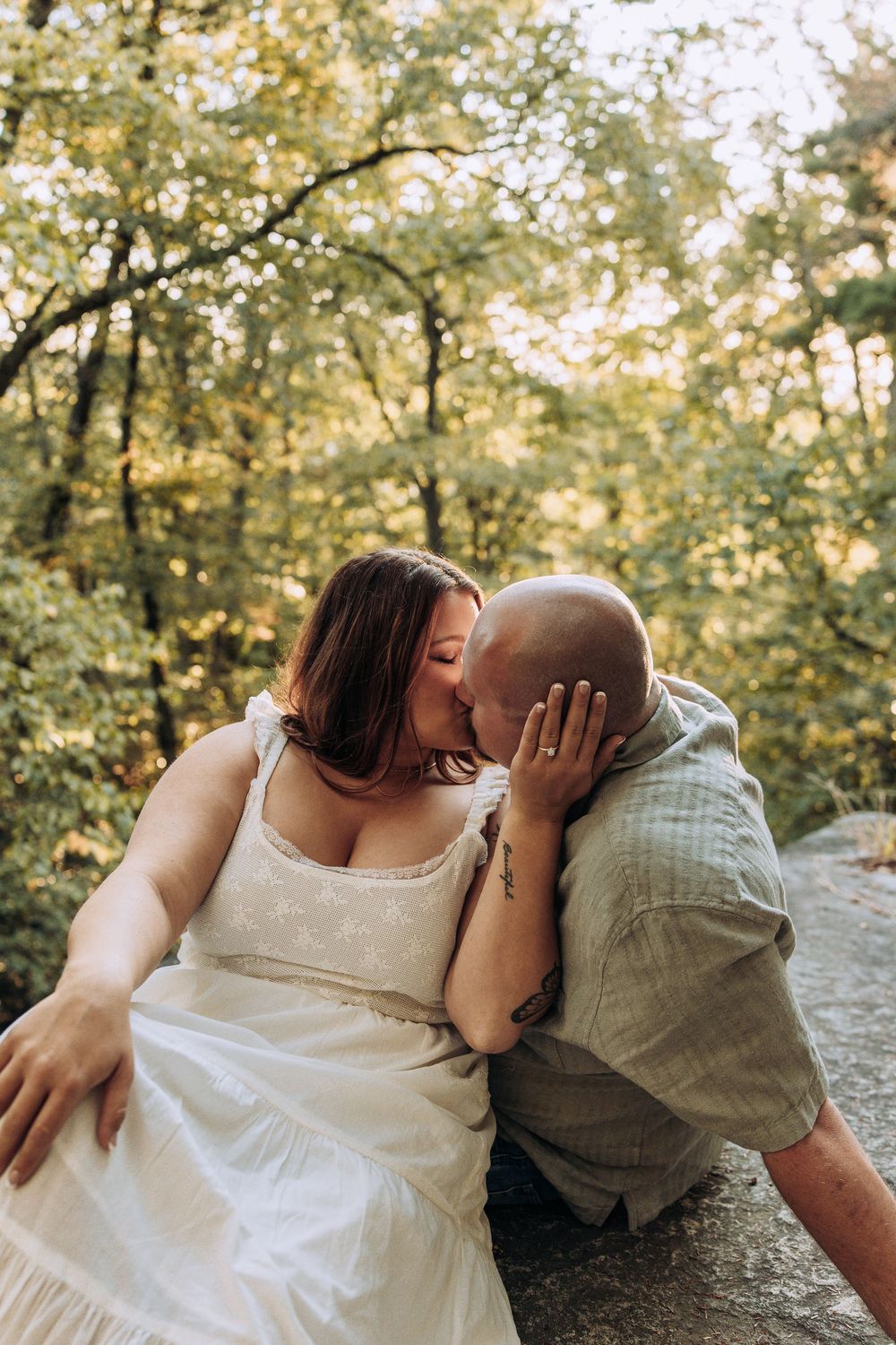Couple sharing a tender kiss on a rocky surface with green trees in the background.
